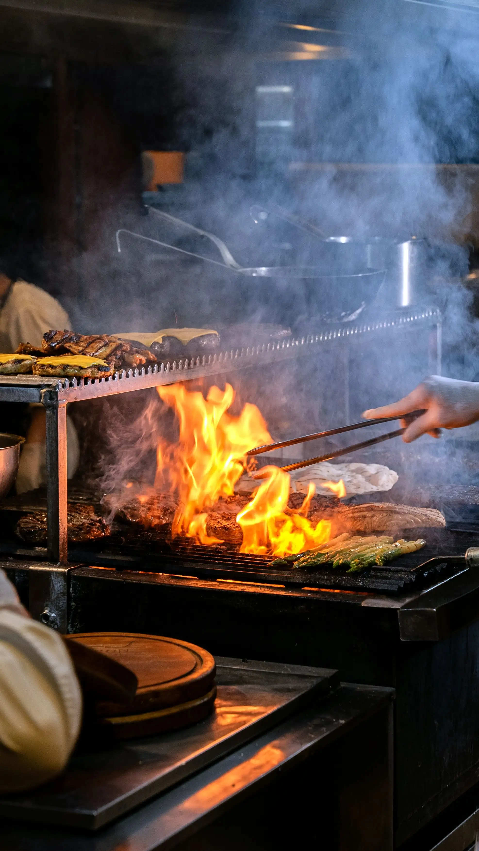 Tongs turning vegetables and meat over the open flame of the grill, smoke rising
