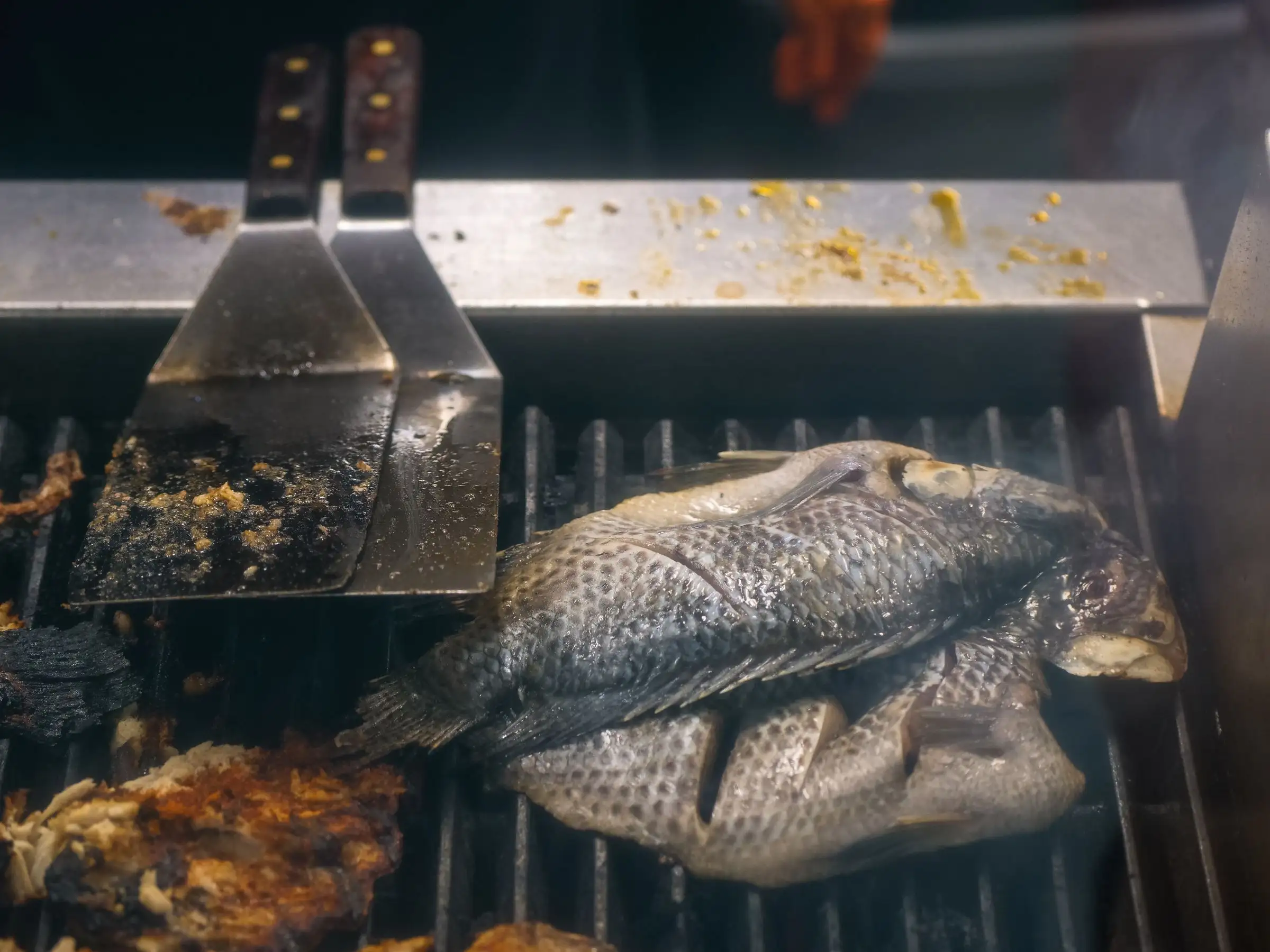 A whole fish crisping on the grill beside a steel spatula